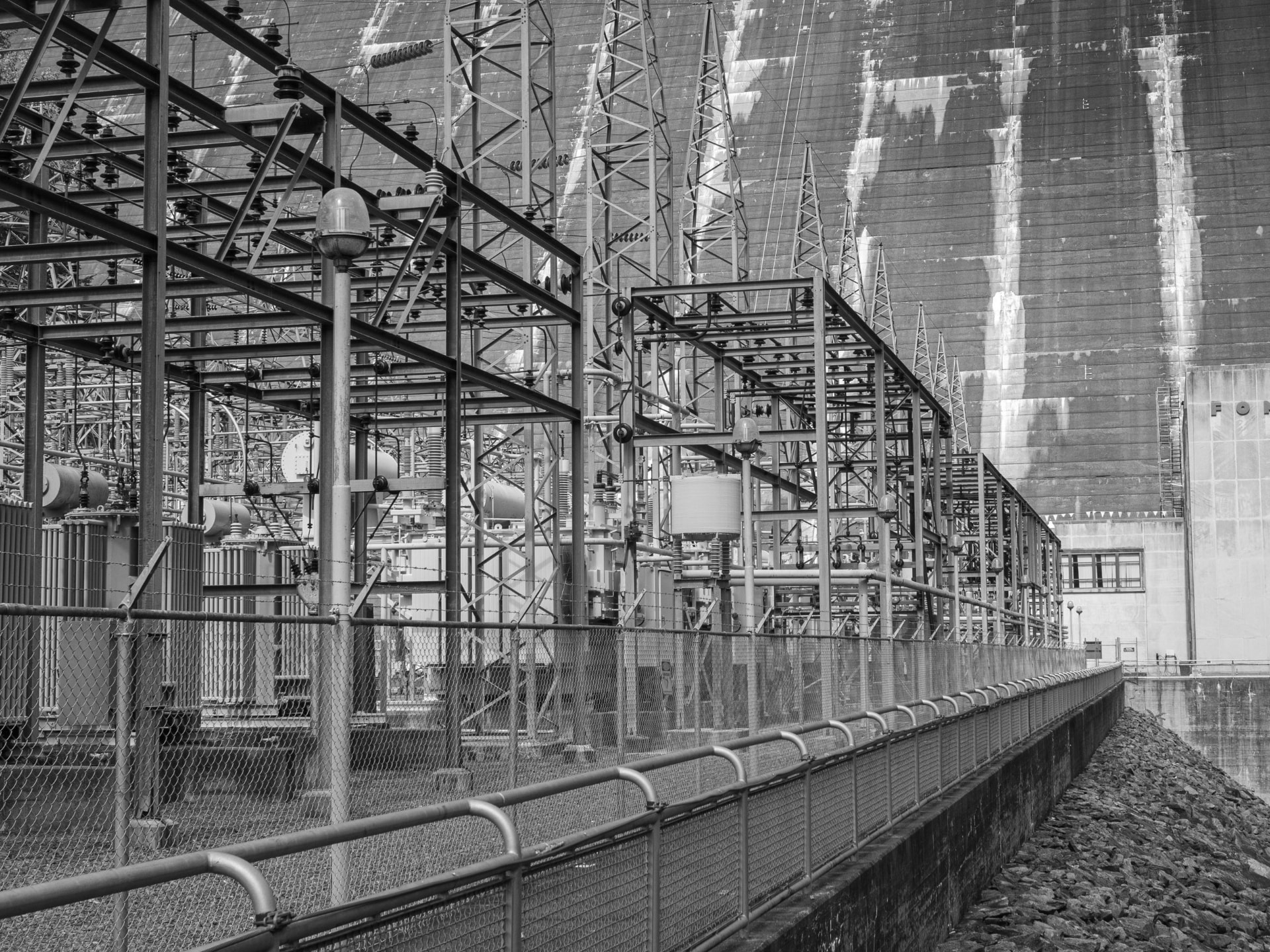 Black and white view of high-voltage switchgear, transformers and transmission towers at a dam power station