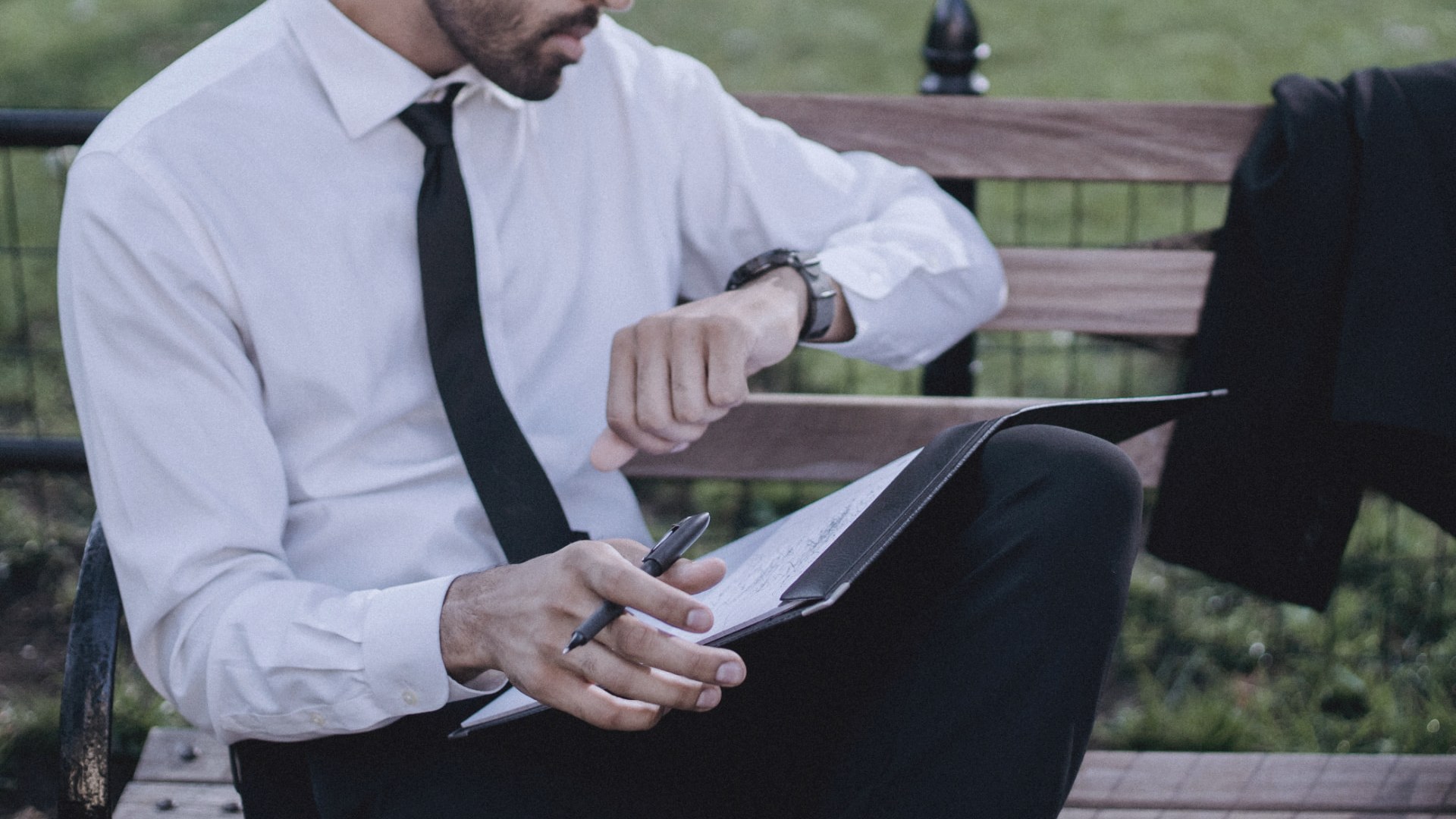 A professional in a white shirt and tie reviewing notes in a folder while checking his watch on a park bench
