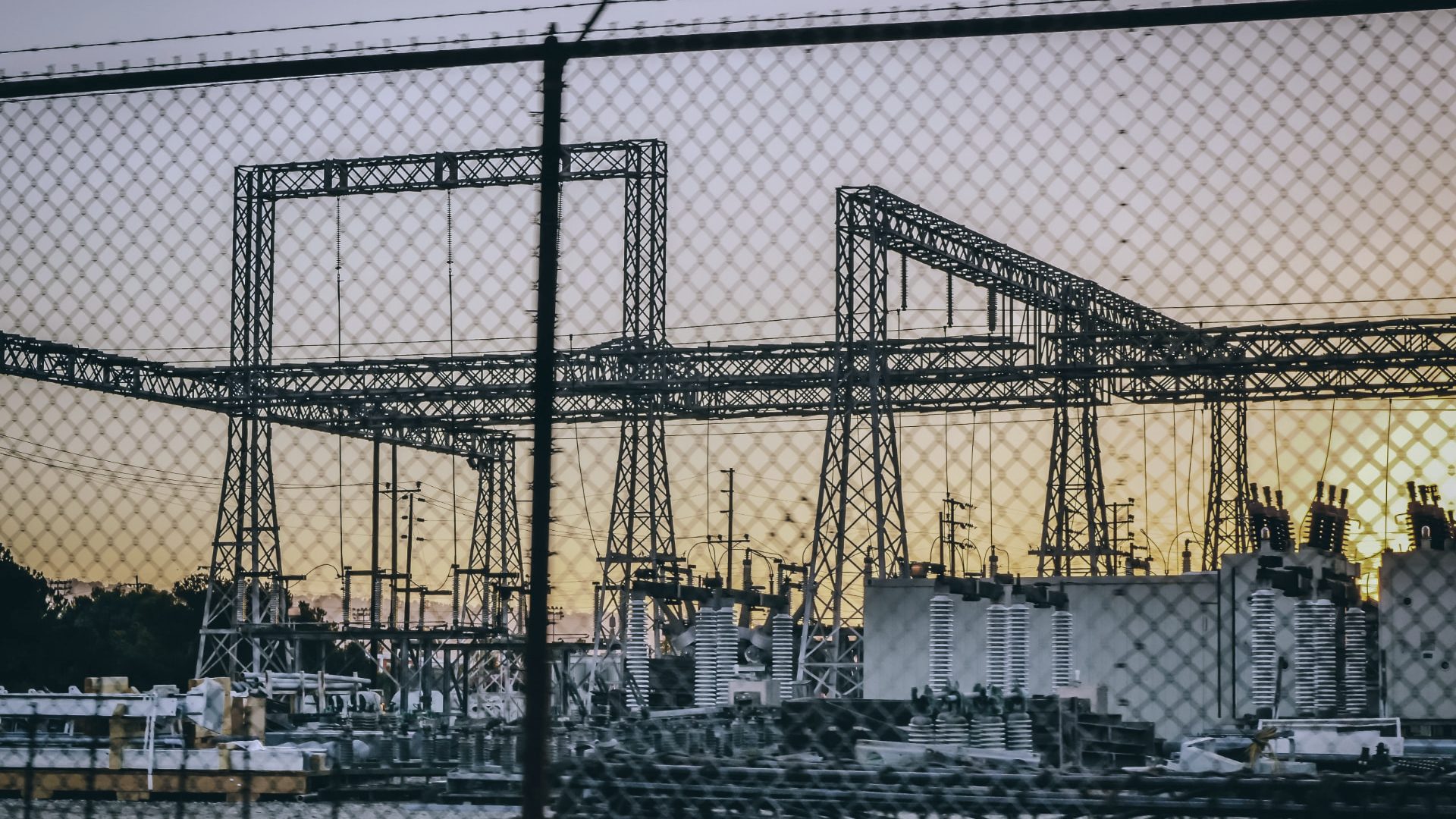 High-voltage substation and transformer yard silhouetted at dusk behind a perimeter fence