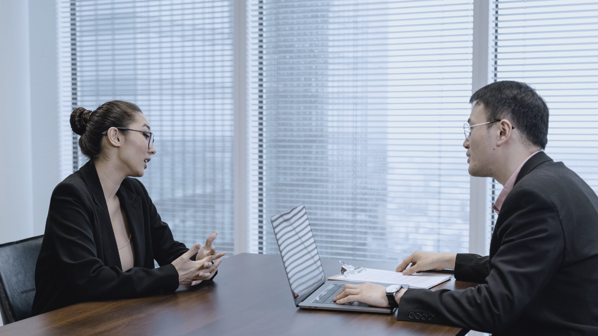 Two people in business attire in a job interview conversation across a table in a bright office