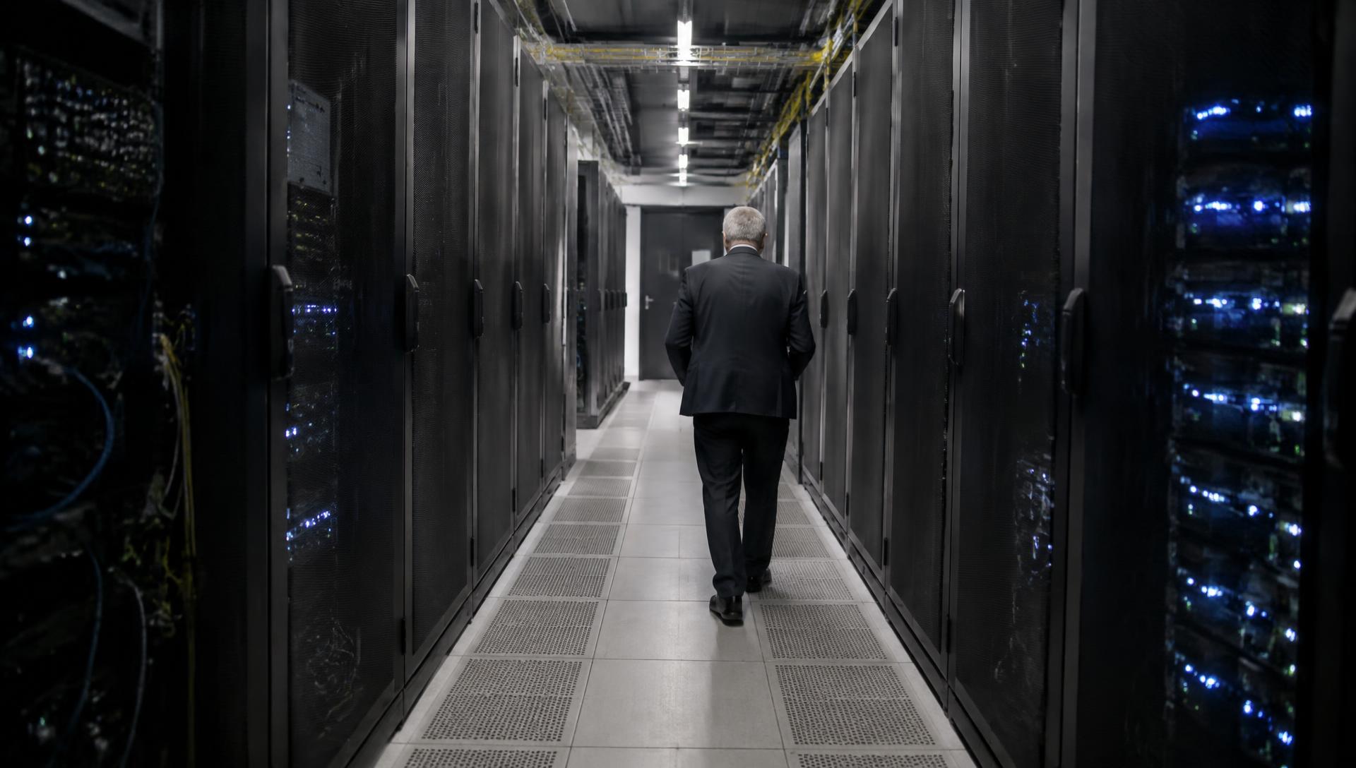 A senior figure walking alone down a long aisle of server racks in a data hall